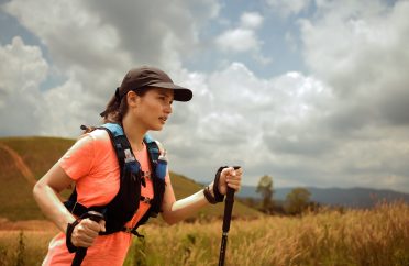 Young women active trail running across a meadow on a grassy trail high in the mountains in the afternoon with trekking pole